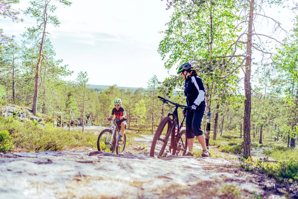 Metsämaisema, jossa nainen on pysähtynyt pyörän viereen katsomaan kun poika ajaa kallioista polkua pitkin ylöspäin.
