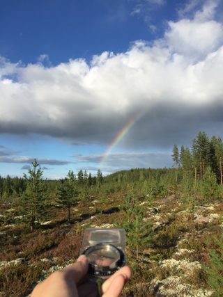 Kuva metsäaukiolta. Kuvaajalla kädessä kompassi joka osoittaa sateenkaarta kohti.